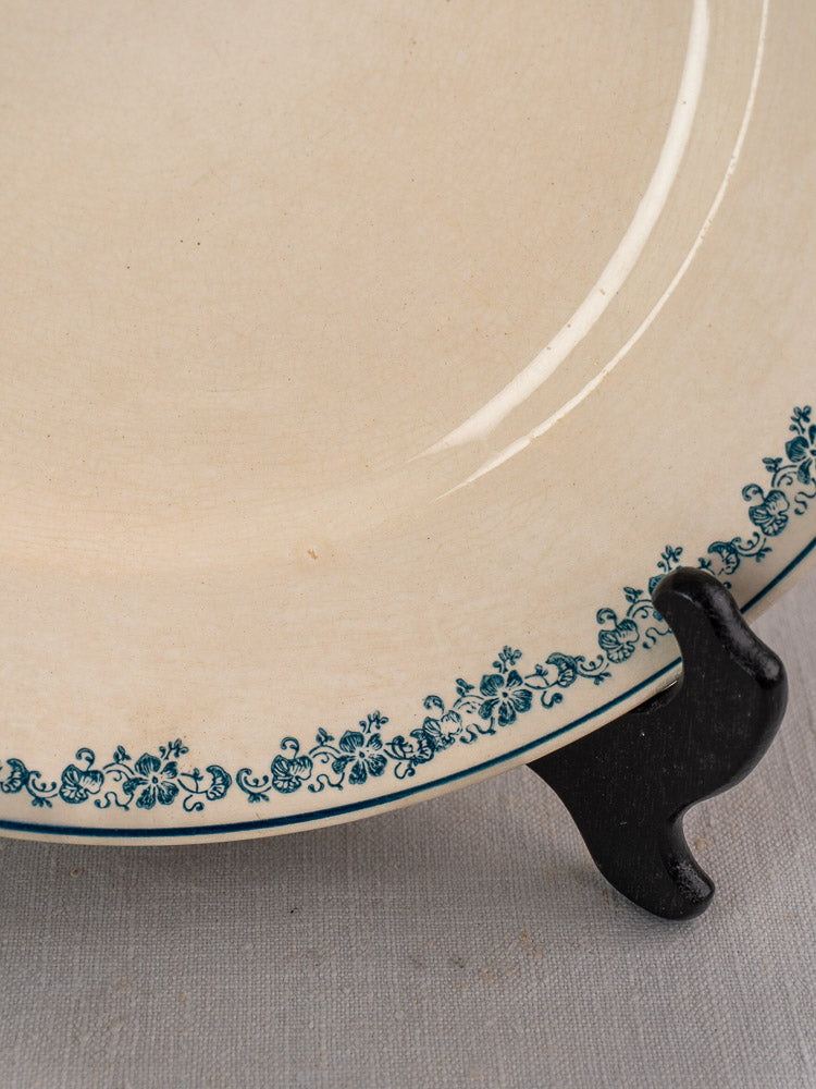 Antique French faience plate, Longwy Capucia pattern with teal floral border on black stand, light gray textured surface.