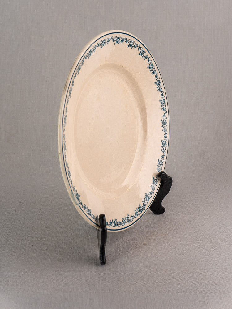 Antique French faience plate, Longwy Capucia pattern, teal floral border, on black stand, gray background.