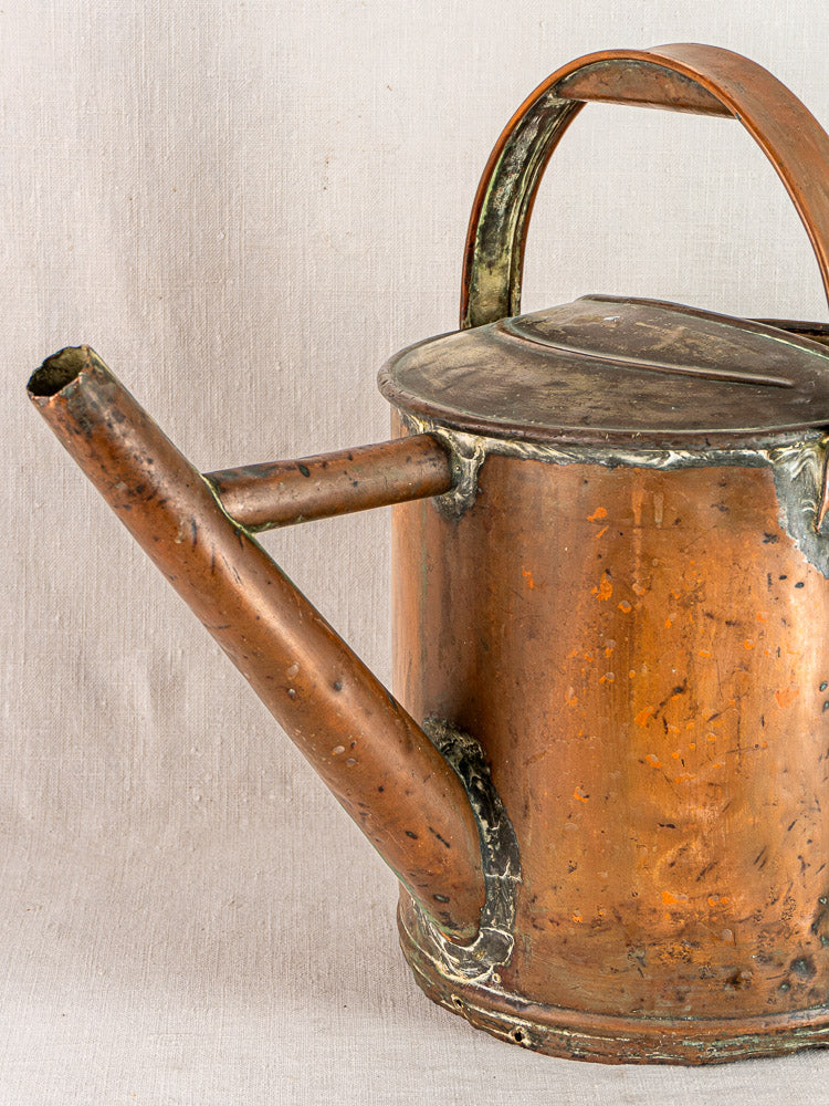 Antique French hand-hammered copper watering can with large spout, curved handle, aged patina on fabric background.