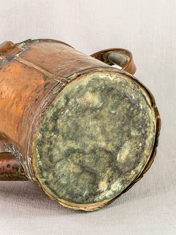 Weathered, tarnished bottom of antique French copper Arrosoir watering can on neutral fabric background.