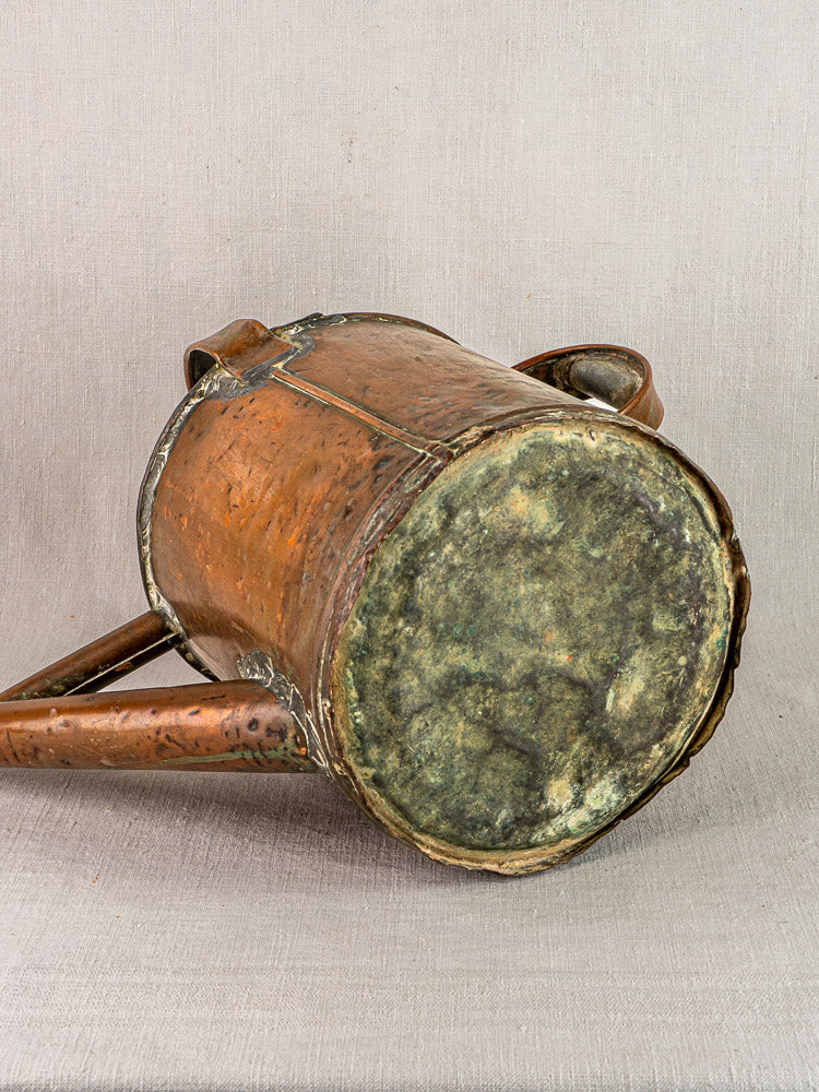 Antique French hand-hammered copper arrosoir watering can with weathered patina, on neutral fabric.