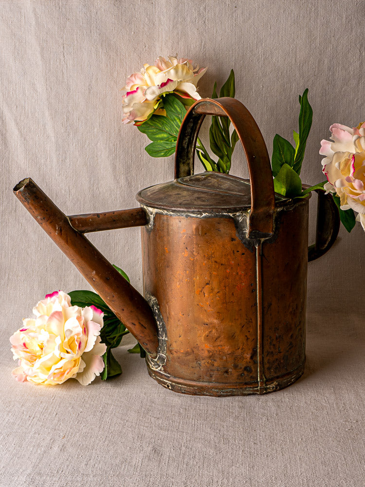 Antique French hand-hammered copper watering can with silk peonies, arranged on neutral fabric.
