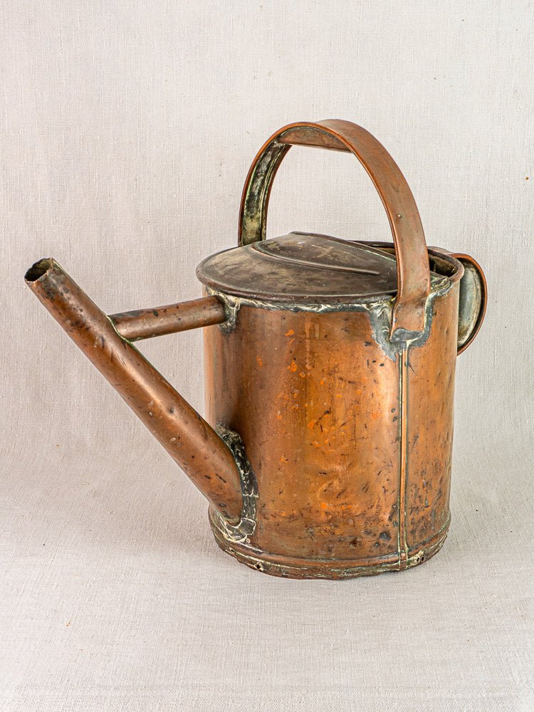 Antique French hand-hammered copper arrosoir with curved handle, resting on a neutral fabric surface.