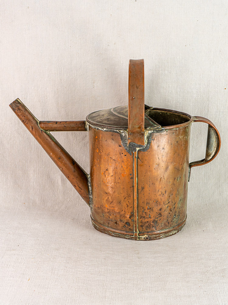 Antique French copper watering can with hand-hammered finish and rich patina on light fabric background.