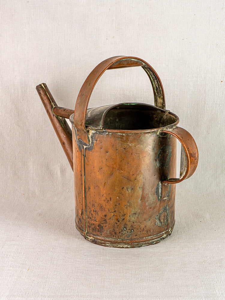 Antique French copper watering can with hand-hammered finish and curved handle on neutral fabric.