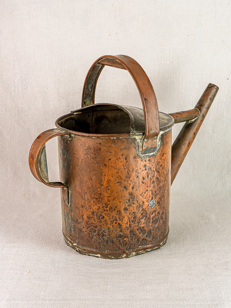 Antique French copper hand-hammered watering can with curved handle and long spout on light fabric.