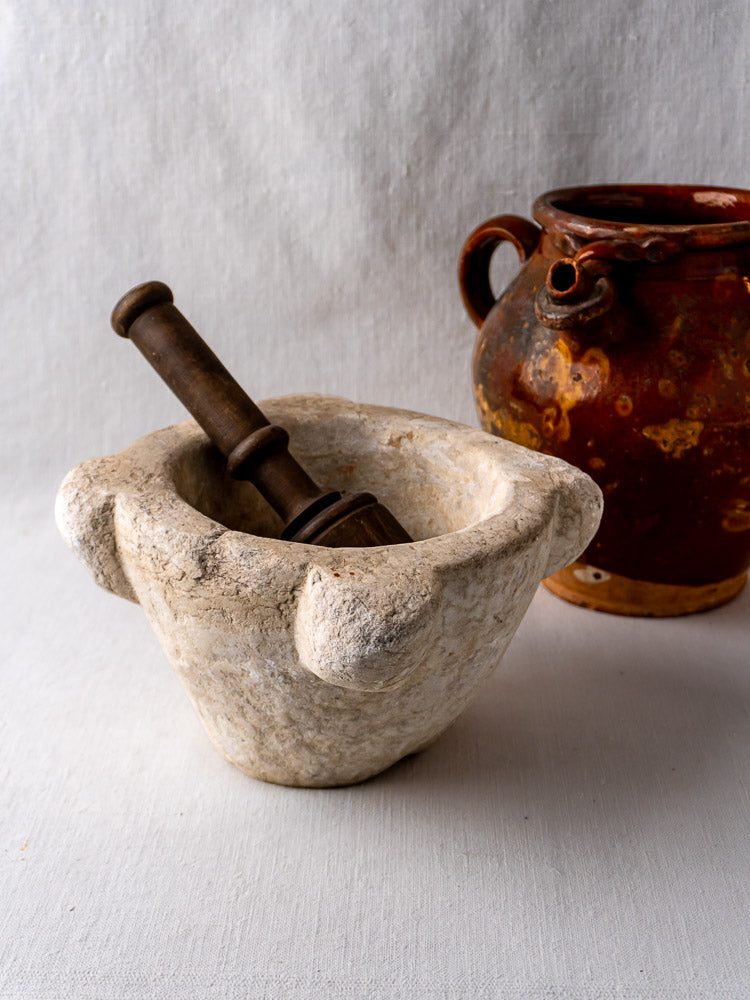 Antique French marble mortar with two handles and dark wooden pestle on light cloth surface.