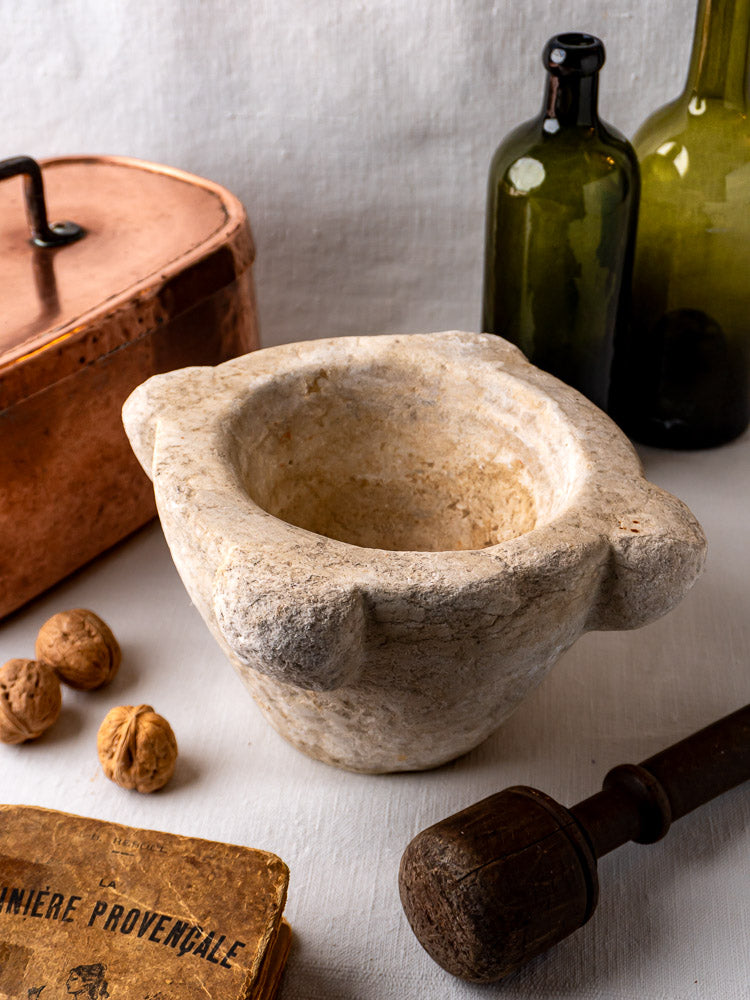 Antique French marble mortar and pestle with walnuts, green bottles, copper pot, and old cookbook on linen.