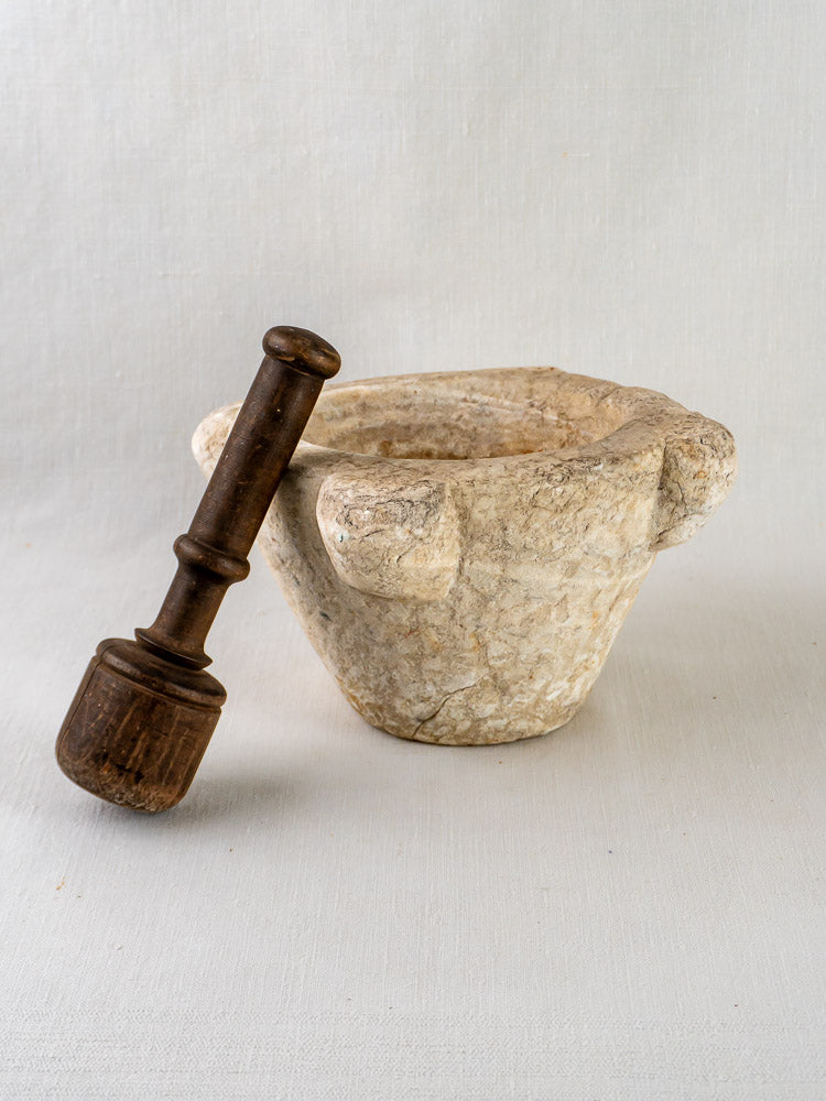 Antique French marble mortar with two handles and dark wooden pestle on light cloth surface.