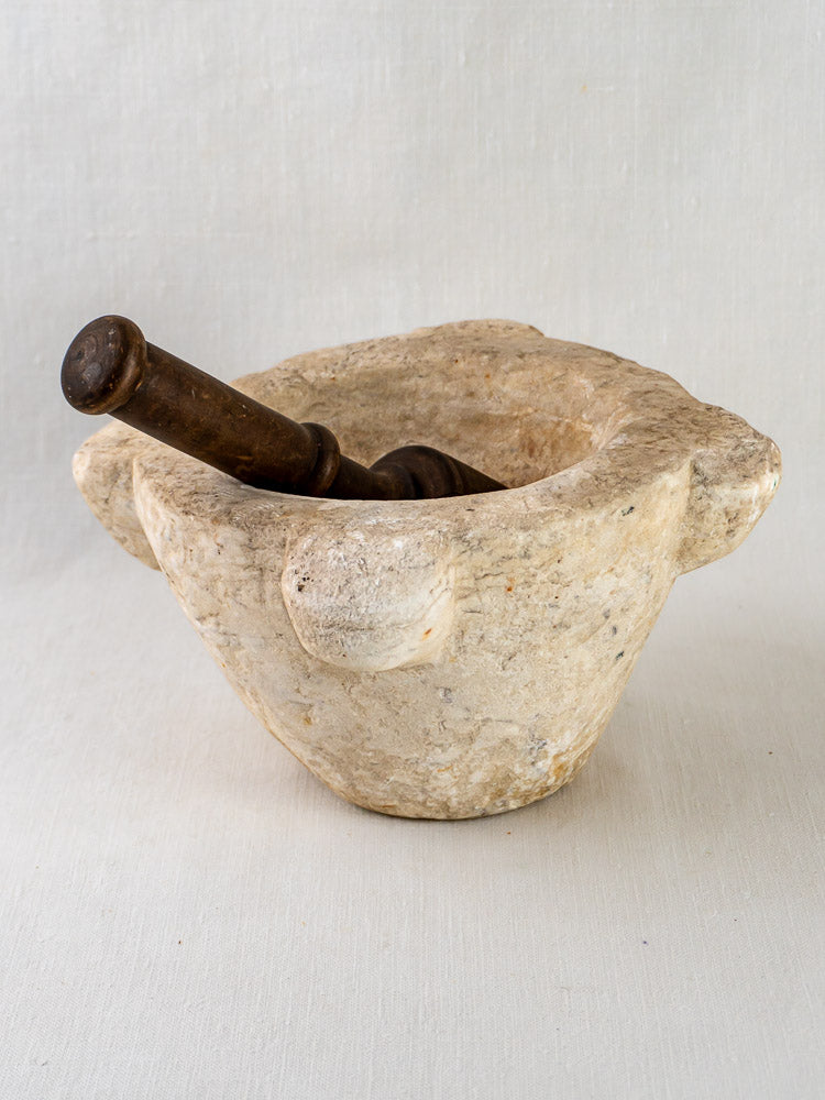 Antique French marble mortar with wooden pestle resting inside, displayed on light fabric.