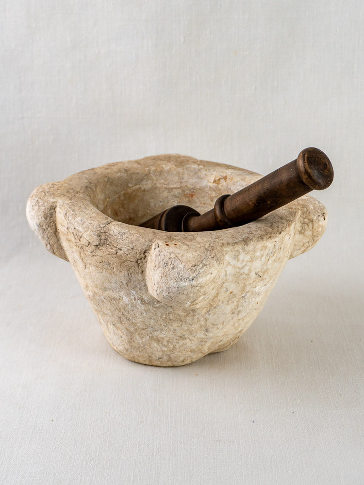Hand-carved marble French mortar with three handles and a dark wooden pestle on a light background.