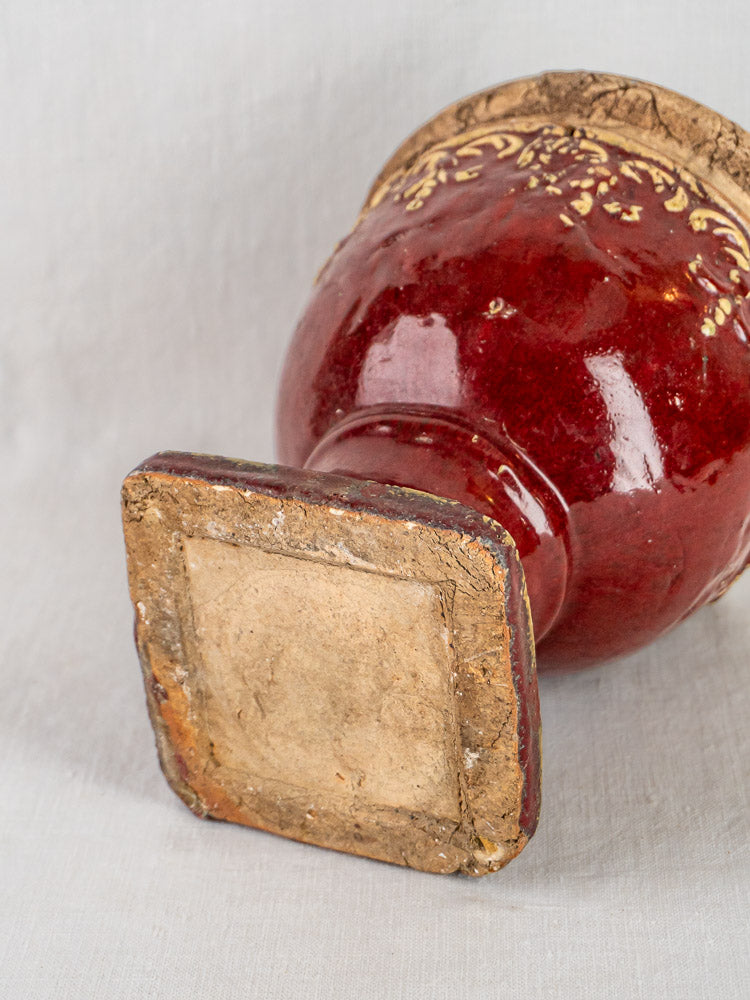 Antique French oxblood terracotta urn with gold accents, worn square base, on neutral fabric.