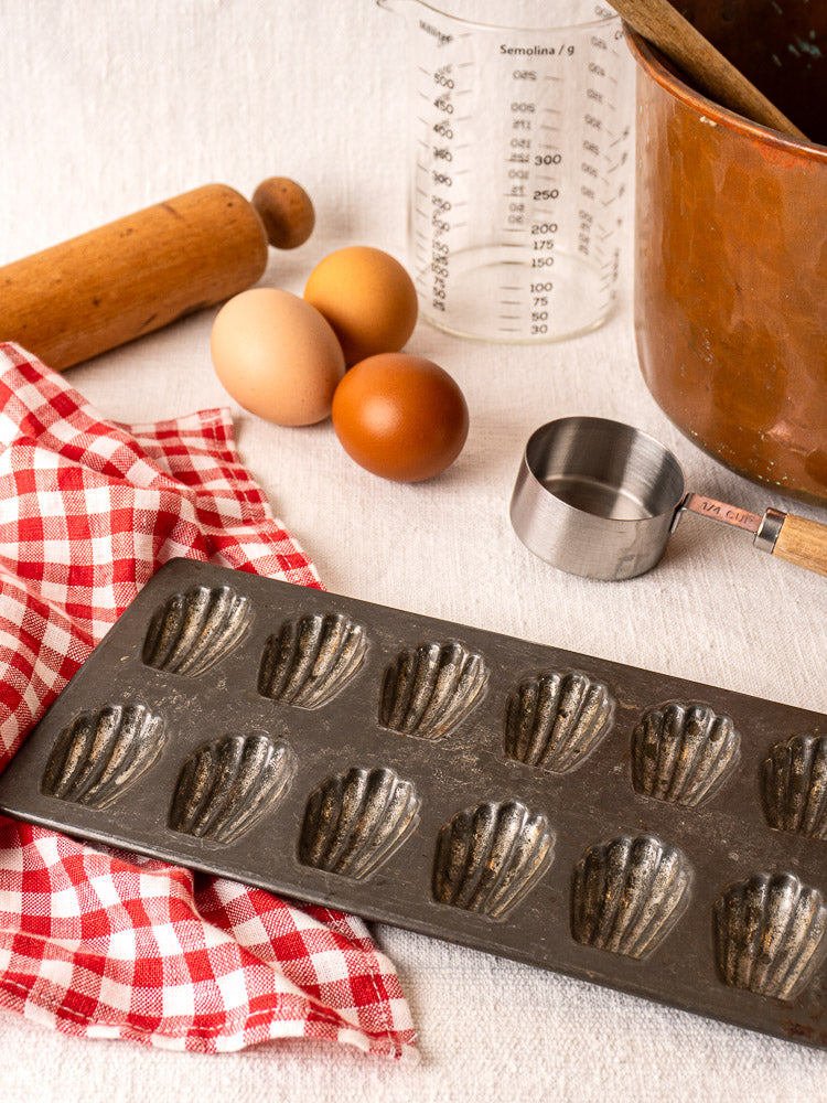 Antique French tinned steel mini madeleine pan with eggs, rolling pin, copper pot, and baking tools on white surface.