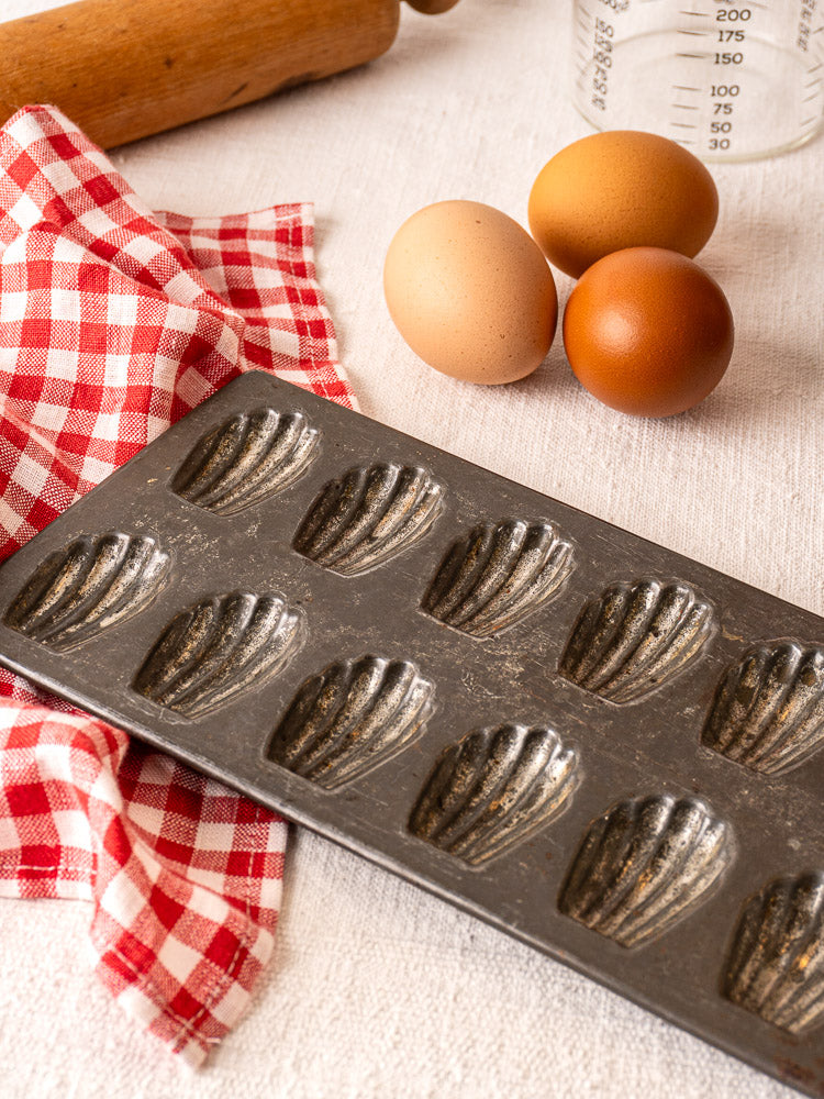 Antique French tinned steel mini madeleine pan with eggs, checkered cloth, rolling pin, and measuring cup.