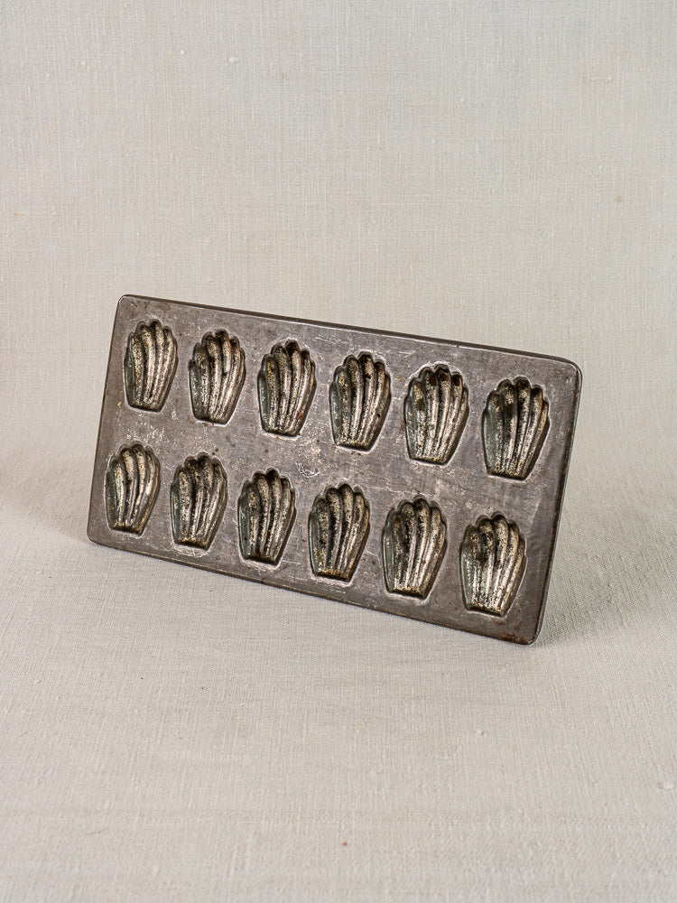 Antique French tinned steel madeleine pan with twelve shell molds on light fabric background.