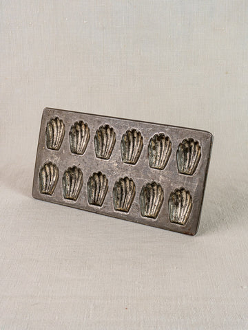 Antique French tinned steel madeleine pan with twelve shell molds on light fabric background.