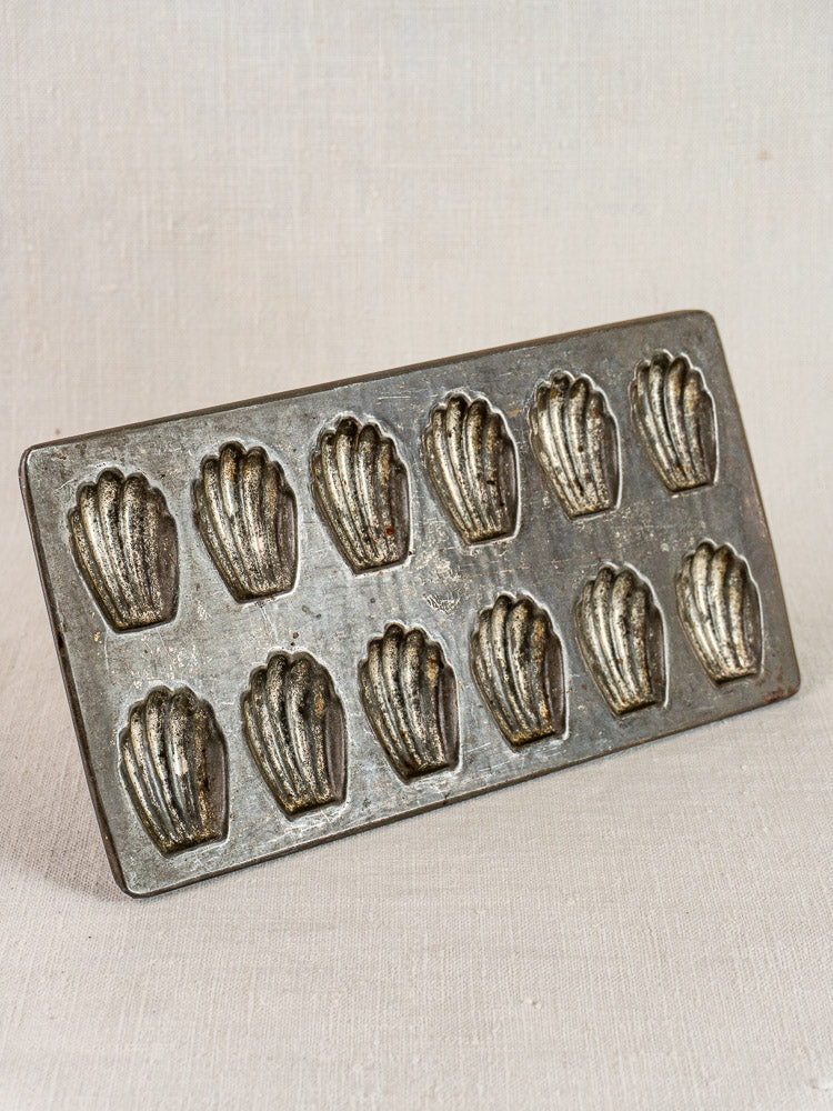 Antique French tinned steel madeleine pan with twelve shell molds against a light, textured backdrop.