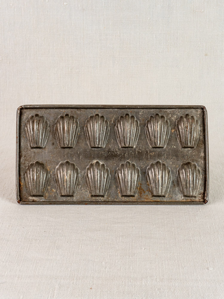 Antique French tinned steel mini madeleine pan with twelve shell-shaped molds on neutral fabric.