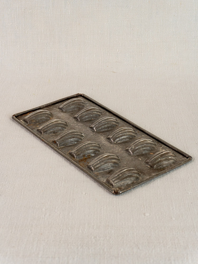 Antique French tinned steel madeleine pan with twelve shell-shaped molds on a neutral textured surface.