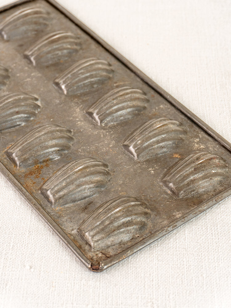 Antique French tinned steel madeleine baking pan with shell-shaped molds on a textured surface.