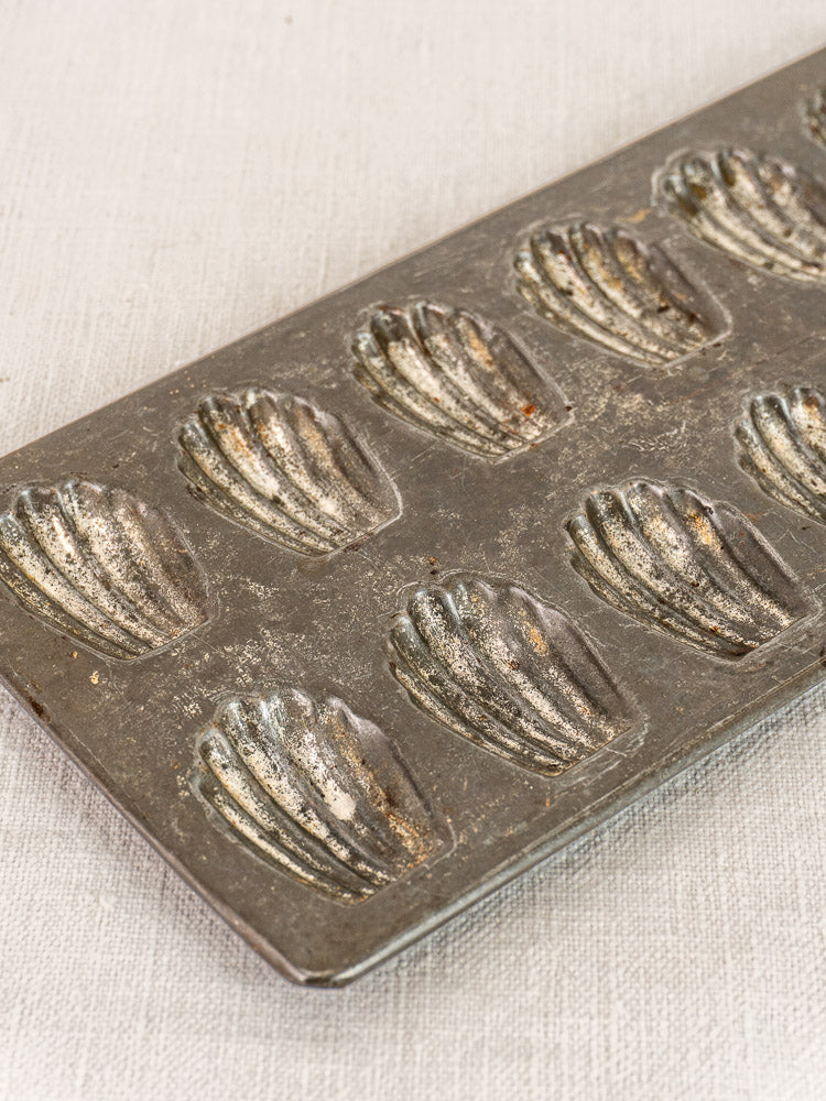 Antique French tinned steel mini madeleine pan with shell designs on a light textured surface.