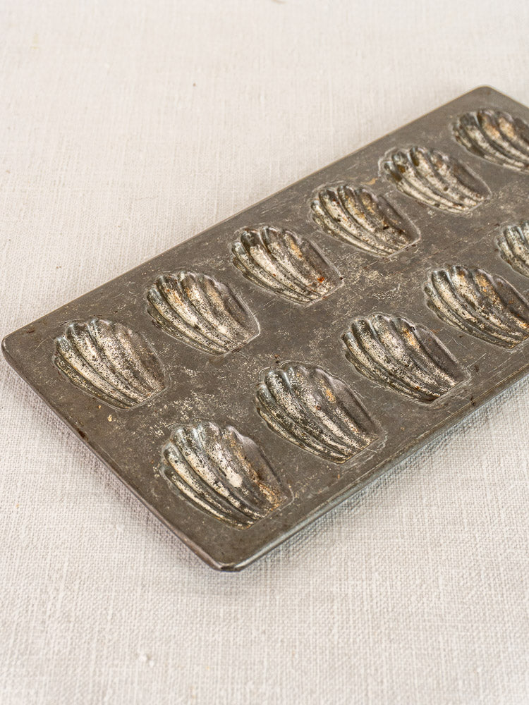 Antique French tinned steel mini madeleine pan with twelve shell molds on textured fabric.