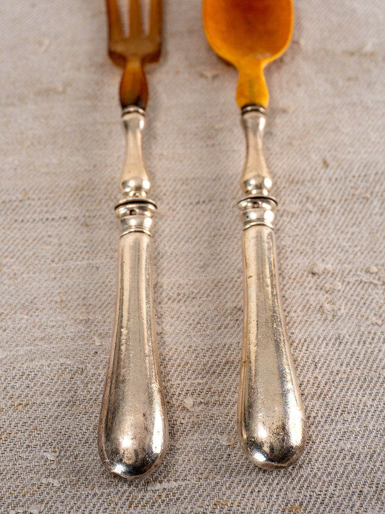 Antique French horn and silver plate salad servers, fork and spoon on beige textured fabric.