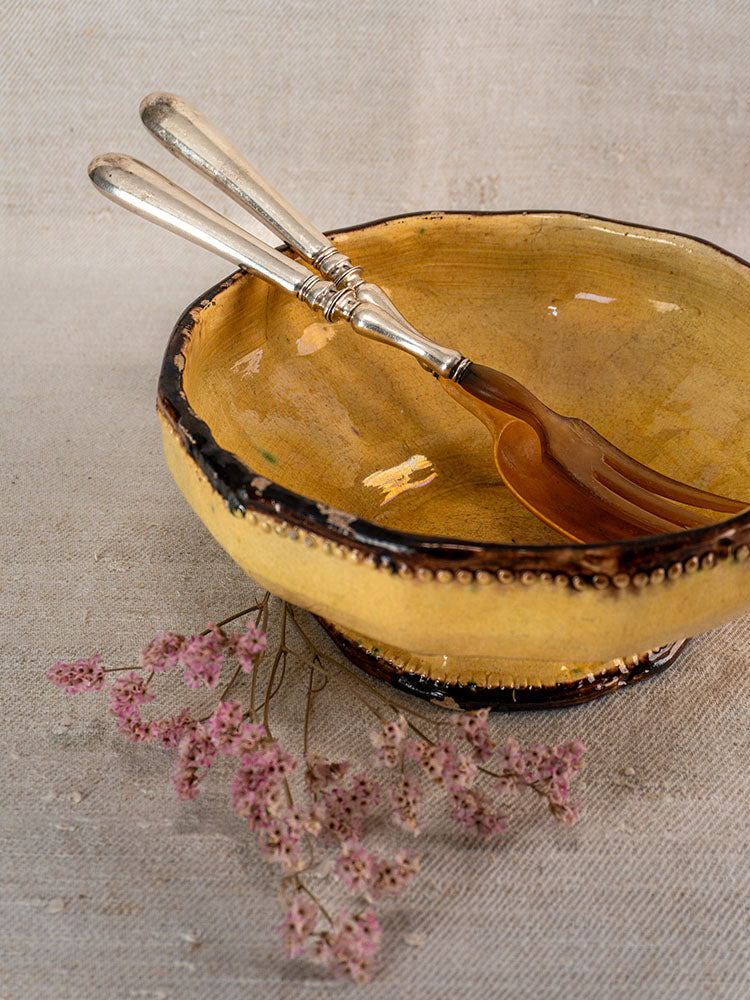 Antique French horn and silver plate salad servers in a yellow ceramic bowl on beige cloth.