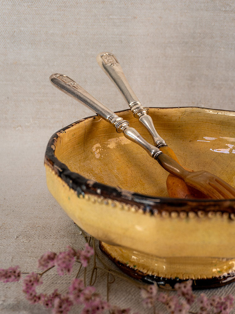 Antique French horn and silver plate salad servers in a yellow ceramic bowl with dark rims.