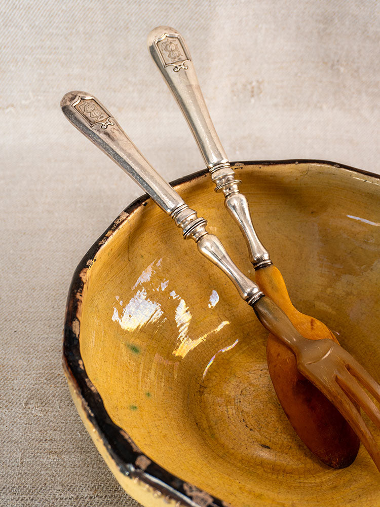 Antique French horn and silver plate salad servers in a rustic yellow ceramic bowl on textured cloth.