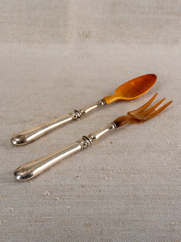 Antique French salad servers with silver-plated handles and brown horn heads on beige textured fabric.