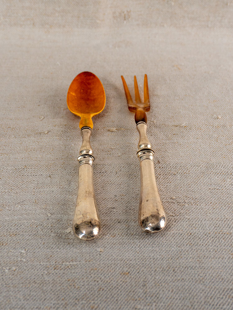Antique French salad servers with amber horn and silver plate, ornate handles on beige textured surface.