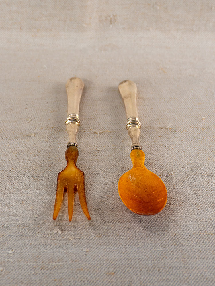 Antique French salad servers with horn tips and silver plate handles on beige fabric background.