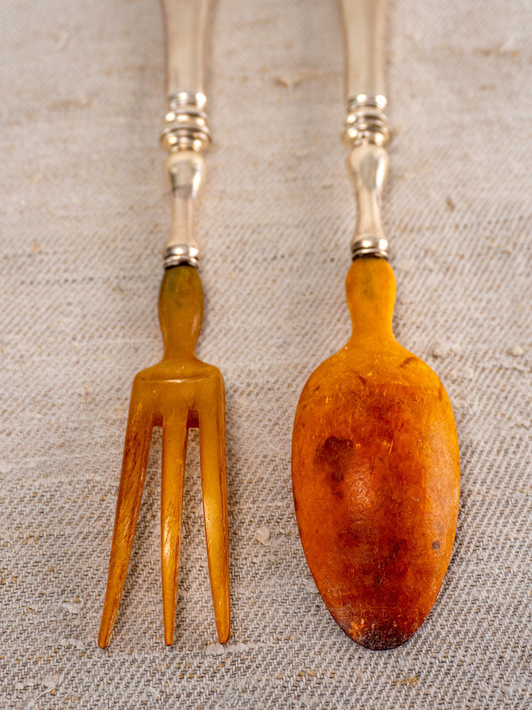 Antique French salad servers with horn ends and silver-plated handles on beige textured fabric.