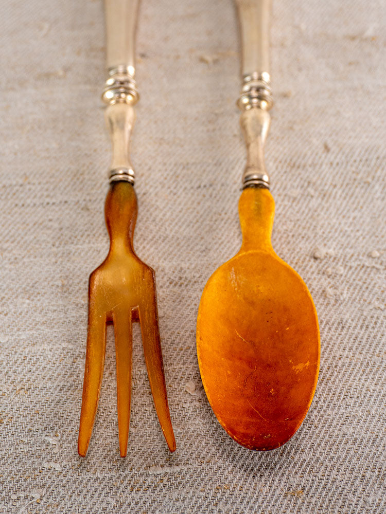 Antique French horn and silver plate salad servers with amber handles on textured beige fabric.