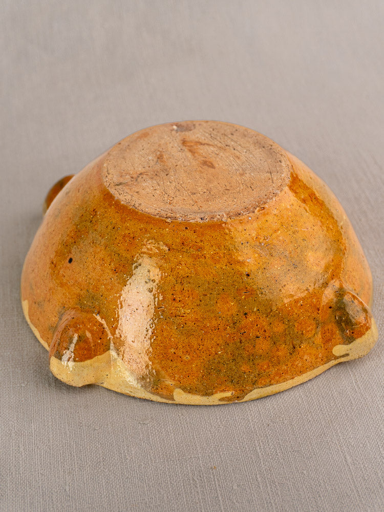 Glossy orange-brown glazed ceramic bowl, upturned to reveal unglazed base and three small feet on gray surface.