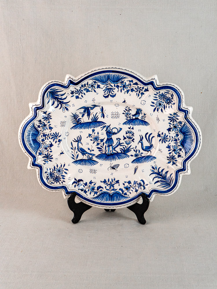 19th-century French faience platter with blue floral and animal motifs on black stand, neutral fabric backdrop.