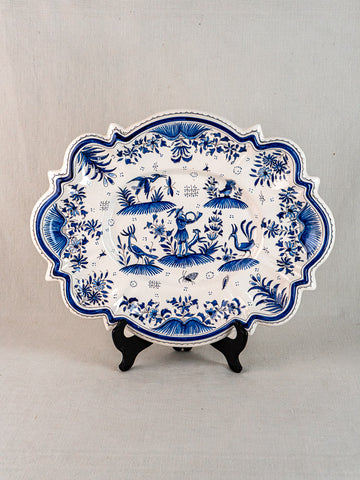19th-century French faience platter with blue floral and animal motifs on black stand, neutral fabric backdrop.