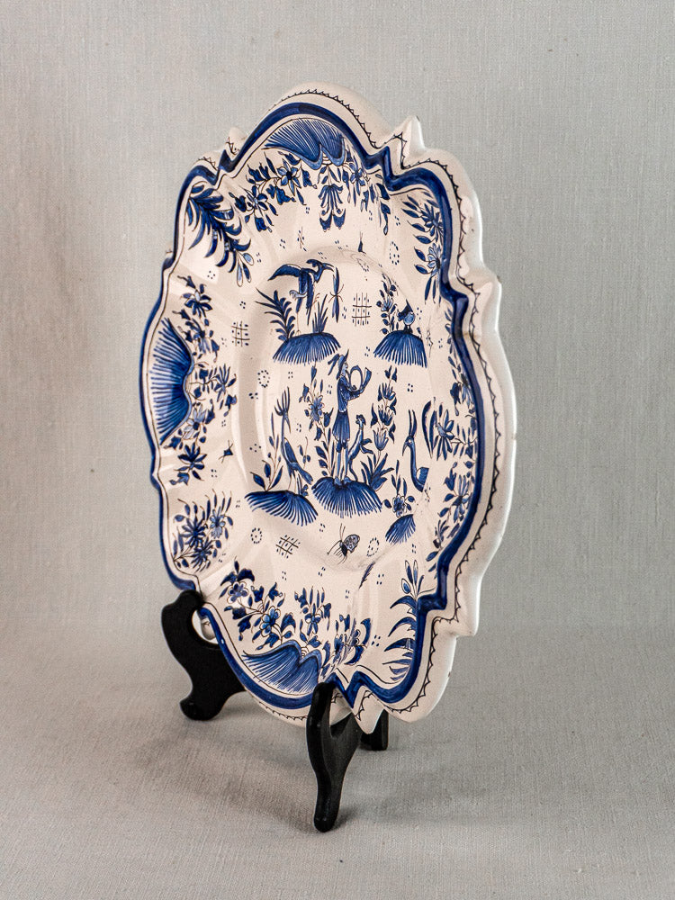 Antique French faience platter with blue floral and landscape motifs on white, upright on black stand.