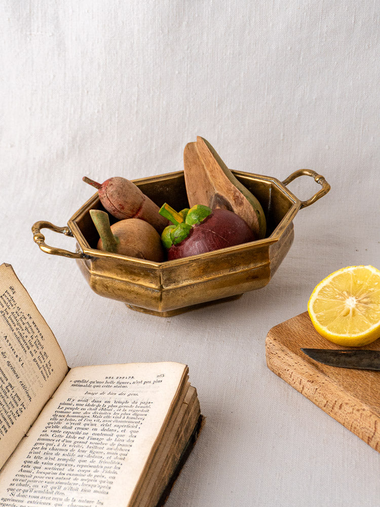 Antique French octagonal brass coupe with wooden fruits and utensils on a light tabletop.