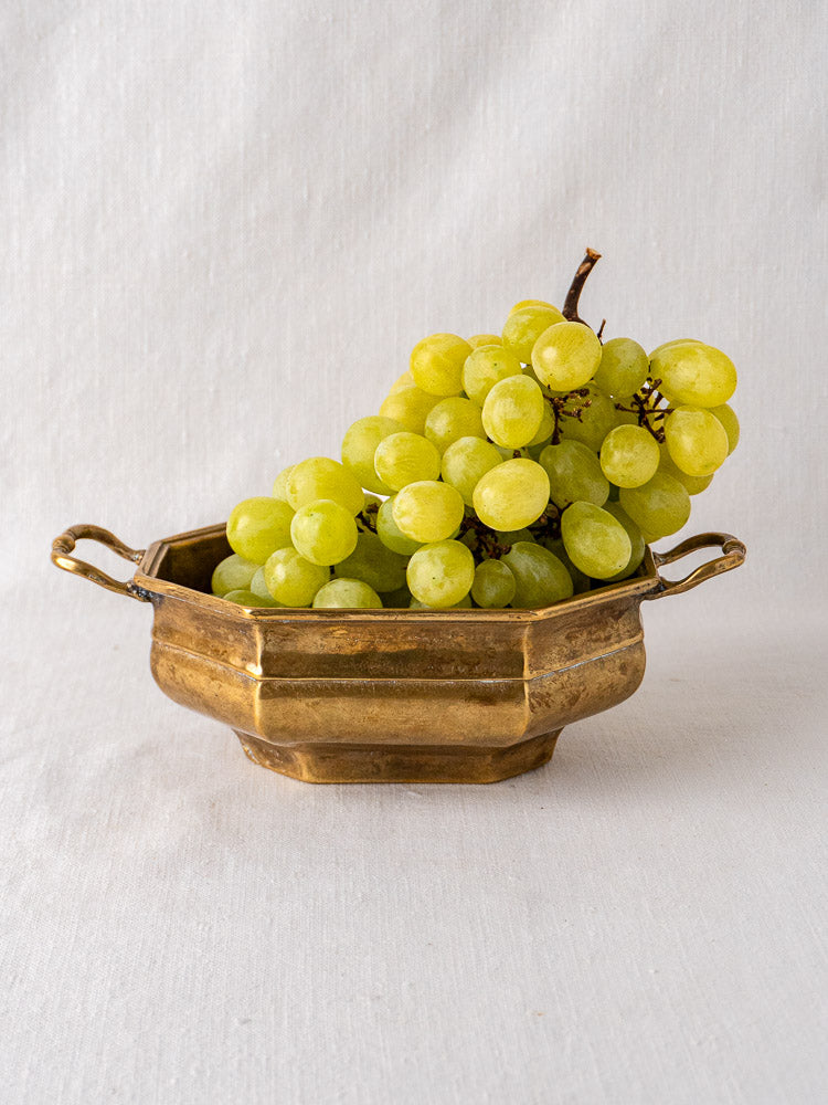 Antique French octagonal brass coupe with two handles, holding green grapes on white fabric.