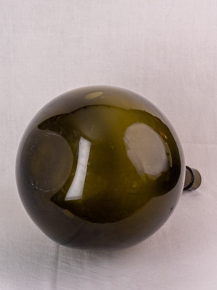 Antique French olive green hand-blown glass demijohn with applied neck ring on a light fabric background.