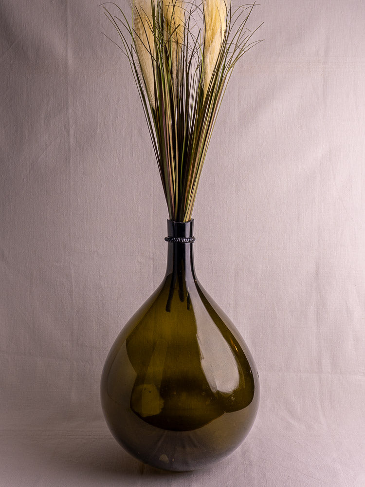 Antique French olive green hand-blown demijohn with applied neck ring, filled with tall dried grass, plain light background.