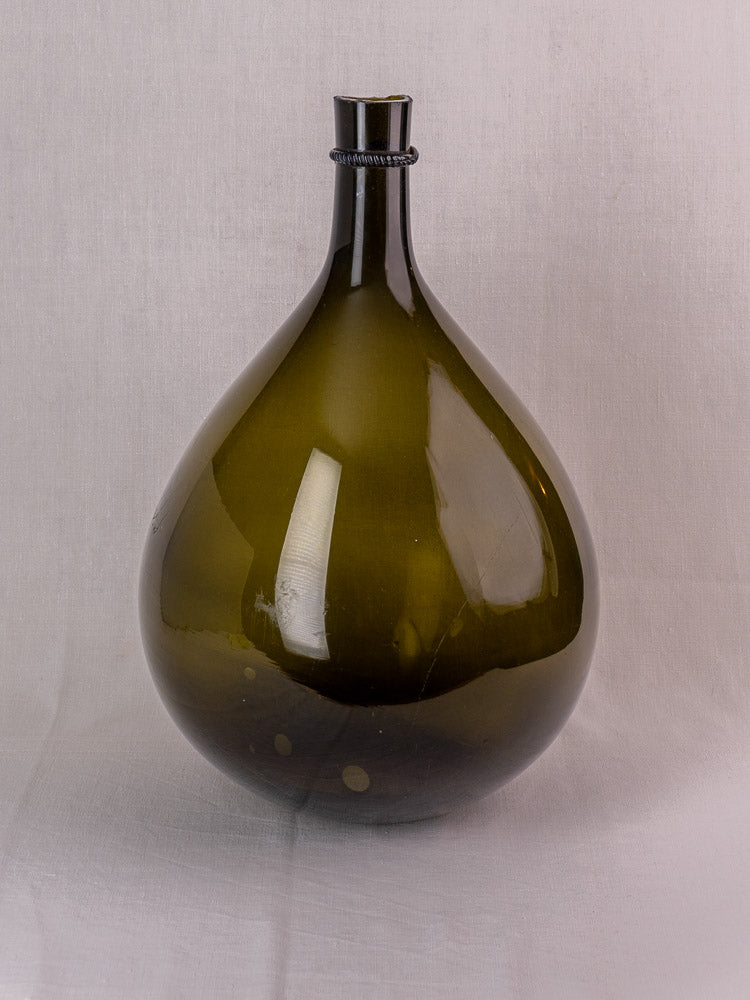 Antique French olive green hand-blown glass demijohn with applied neck ring on light gray textured surface.