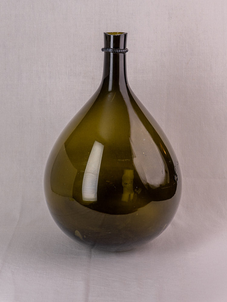 Antique French Demijohn in olive green hand-blown glass, bulbous base, applied neck ring, on light gray background.