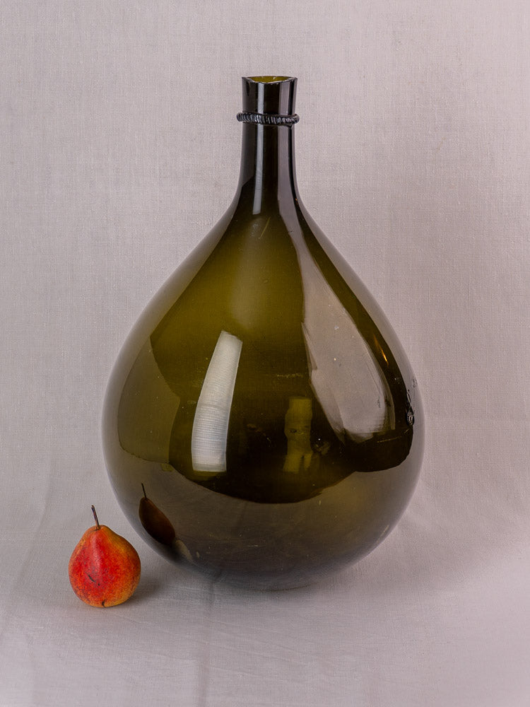 Antique French olive green hand blown demijohn with applied neck ring beside a red pear on light surface.