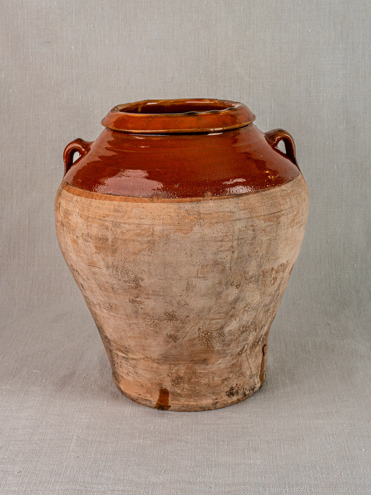 Antique French olive oil jar in glazed terracotta, brown and beige, with side handles, on gray background.