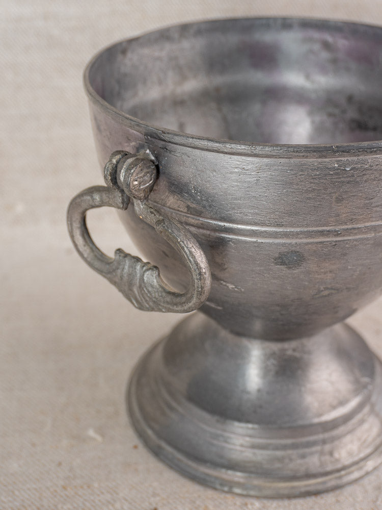 Antique French pewter footed bowl with ornate handle and sturdy base on a textured beige surface.