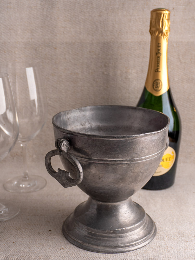 Antique French pewter footed bowl, mid-19th century, displayed with champagne bottle and wine glasses on beige surface.