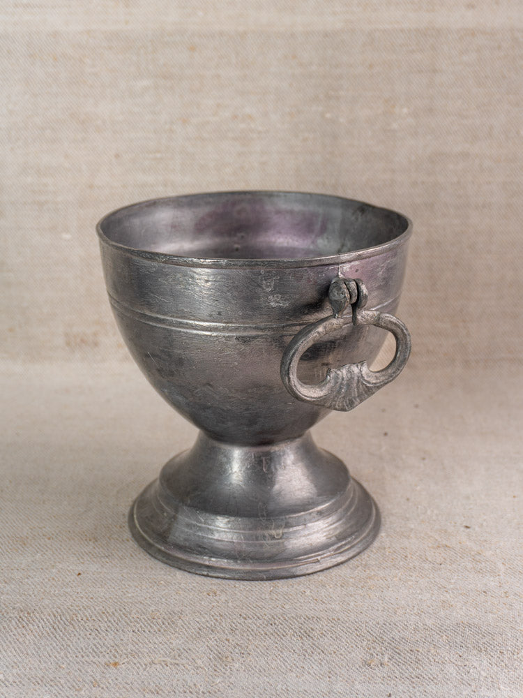 Antique French pewter footed bowl with wide base and ring handle on textured beige fabric background.